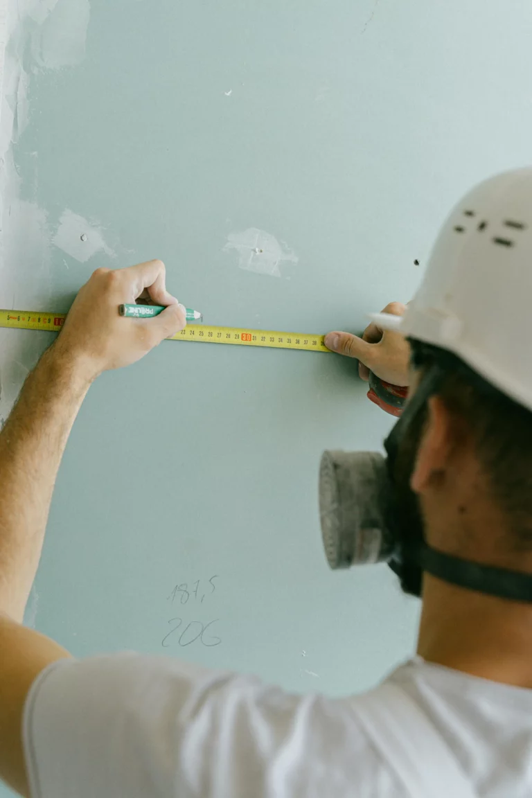 Artisan en casque de chantier mesurant un mur avec un mètre ruban et marquant des repères au crayon sur une plaque de plâtre verte.