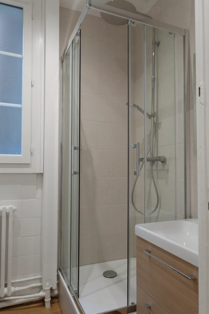 Salle de bain rénovée avec douche à paroi vitrée et carrelage beige.